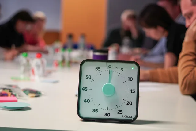 Timer on a table with blurred people in an office meeting setting, emphasizing focus. Arbejdstid, overarbejde og hviletid i danske virksomheder
