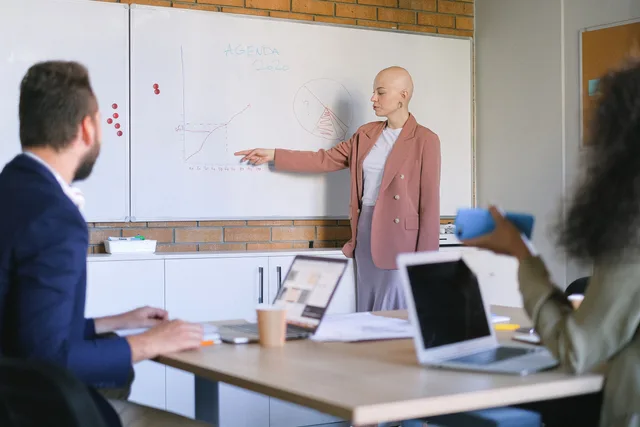 Female manager demonstrating graphs on whiteboard to anonymous coworkers at table with laptops in conference room during meeting. Trivsel på arbejdspladsen: Fra APV til forebyggelse
